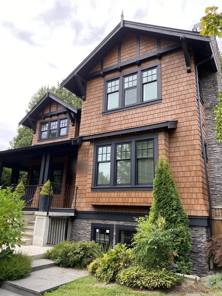 A craftsman house in North Vancouver: cedar shingles above, stone at the base, and every timber element — gable beams, brackets, window frames, porch posts and railings — carried in a near-black. The dark work is what holds the front together, and it is also the work that takes the most weather.