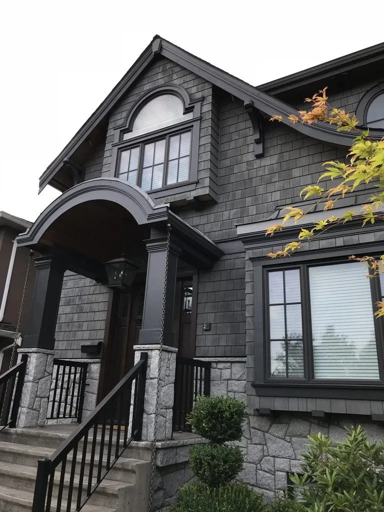Slate-grey shingle walls with white window sashes behind dark frames, an arched porch in the same deep grey, and granite at the base. A near-monochrome front where the only contrast is in the glazing bars — which means every edge has to be cut clean.